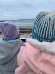 Person wearing a pink coat and blue beanie and the other person wearing a violet beanie with a blurred beach background