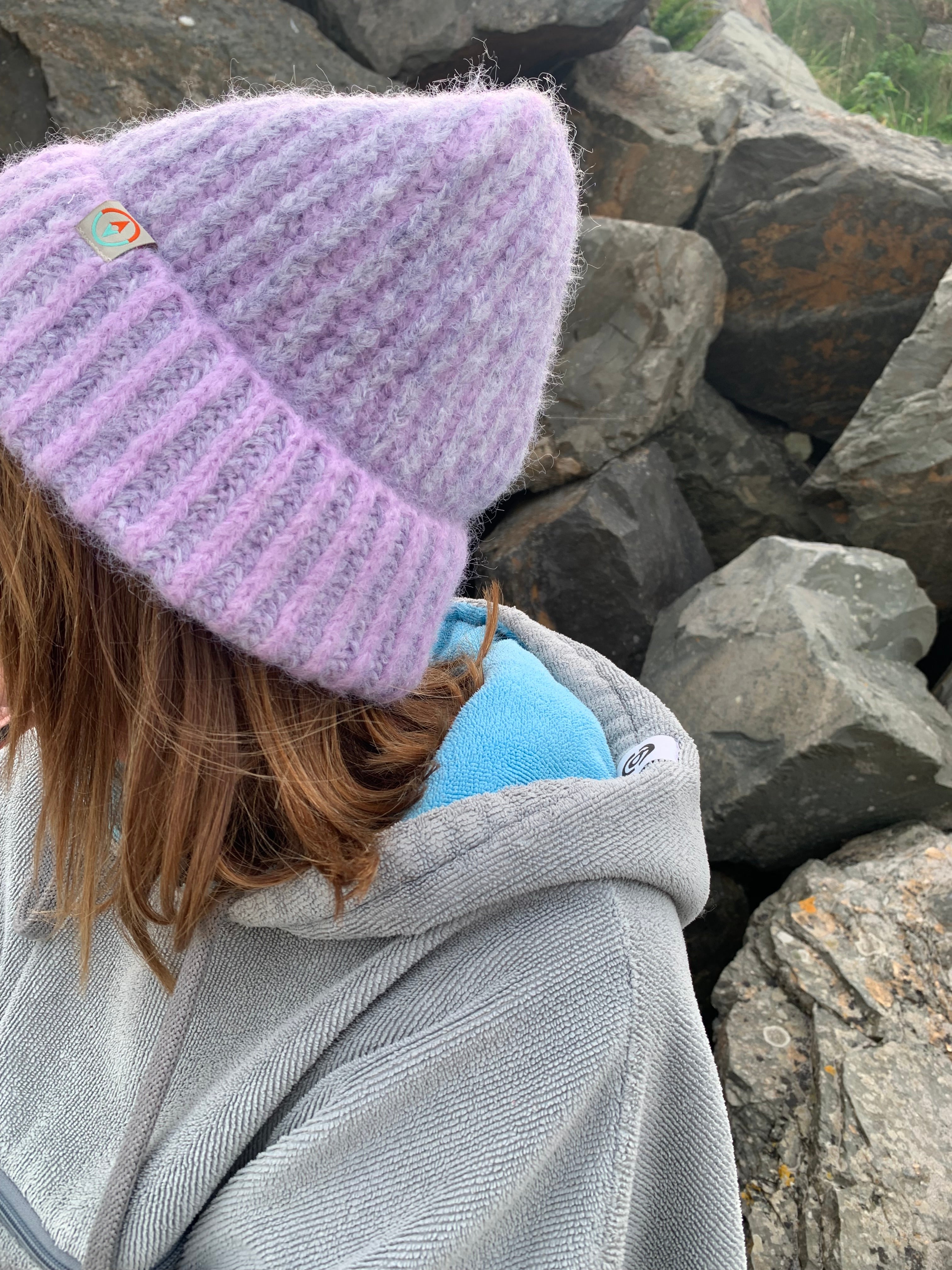 Close-up of a person wearing a gray jacket and purple knit hat with a fluffy texture, standing against a rocky background.