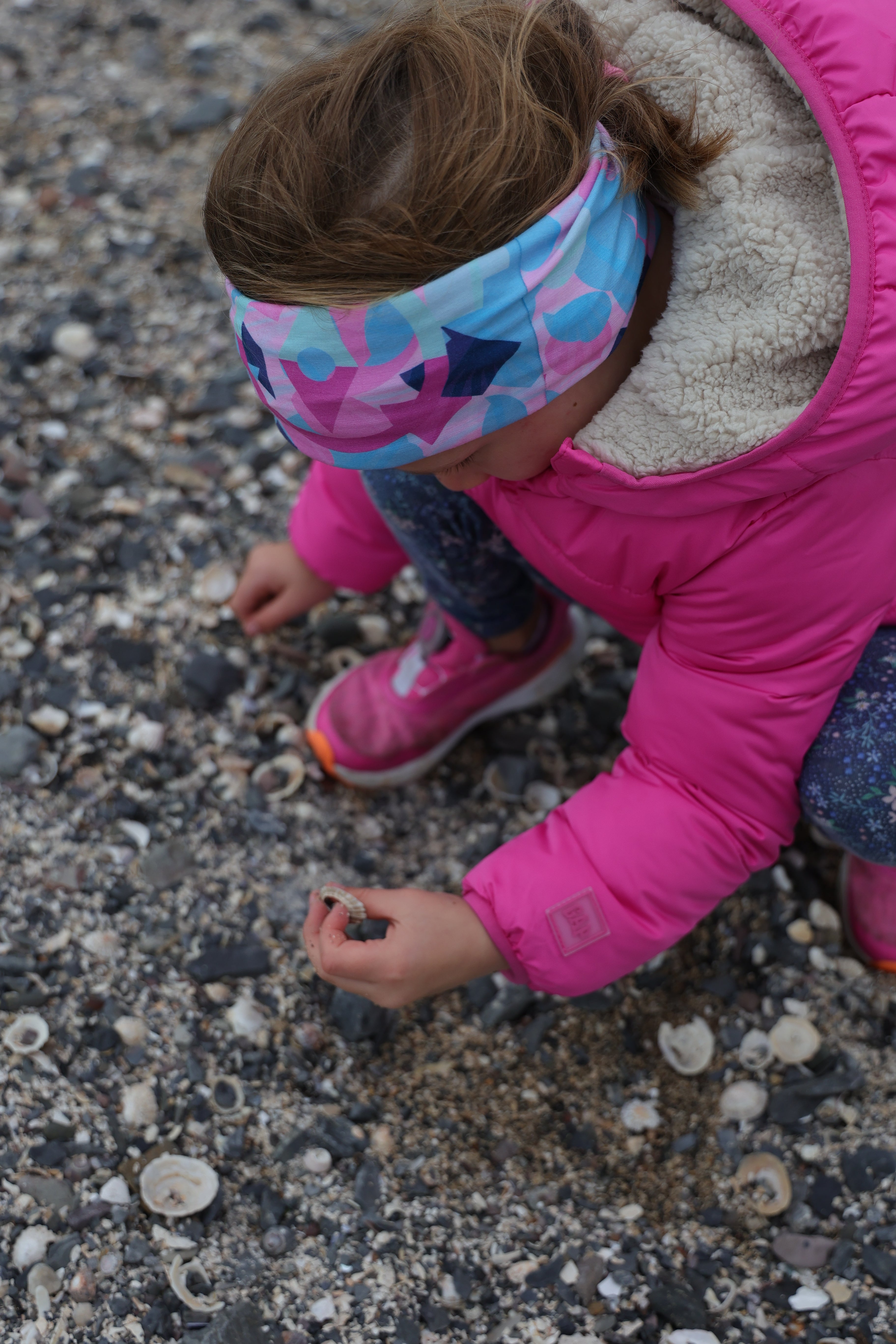 Young girl in a pink coat wearing the Beach Bounty Infinity Band in pinks, blues and navy as she kneels down to collect shells at the beach.