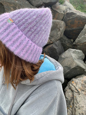 Close-up of a person wearing a gray jacket and purple knit hat with a fluffy texture, standing against a rocky background.