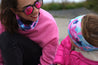 Mother and daughter both wearing the same Infinity Band, mum wearing it as a neck warmer or snood with a pink sweatshirt and pink mirror sunglasses and the young girl wearing the Infinity Band as a headband keeping her ears warm along with a pink coat as they play at the beach.