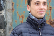 Young man wearing the multiway Infinity Band (snood) around his neck outdoors standing before a coastal boathouse showing its grey and black design inspired by The Giants Causeway in Northern Ireland. 