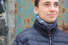 Young man wearing the multiway Infinity Band (snood) around his neck outdoors standing before a coastal boathouse showing its grey and black design inspired by The Giants Causeway in Northern Ireland. 