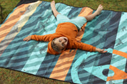 Sustainable waterproof picnic blanket made from recycled polyester, oversized yet packable as it folds flat into a laptop size to slide into a backpack. Zip corner pocket and stake loops and sand pockets to keep it in place. 